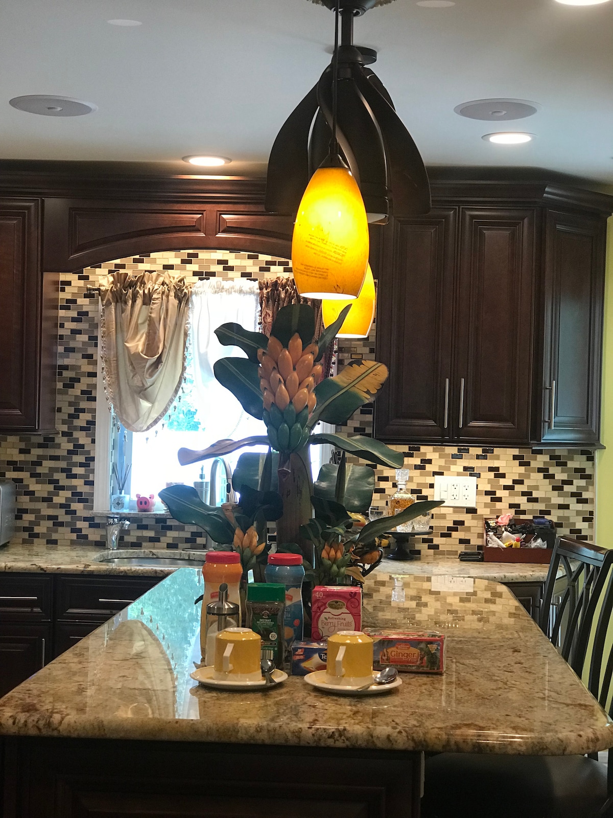 A well-appointed kitchen features a large granite island topped with decorative items and two yellow cups. A striking light fixture hangs above, complemented by a lush floral arrangement. The mosaic tile backsplash adds texture, while natural light enters through the window, framed by soft drapes.