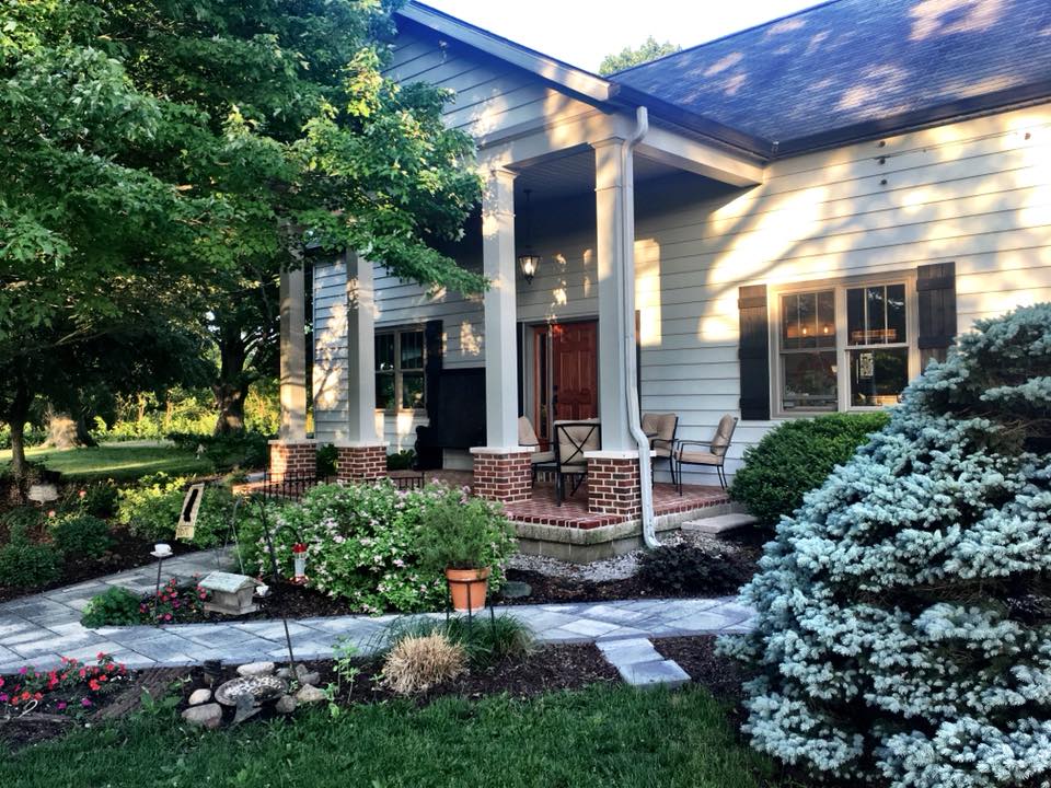 The entrance features a spacious porch adorned with two chairs and a welcoming door. A stone pathway leads through a landscaped garden with a variety of plants, including shrubs and flowers, under the shade of mature trees.
