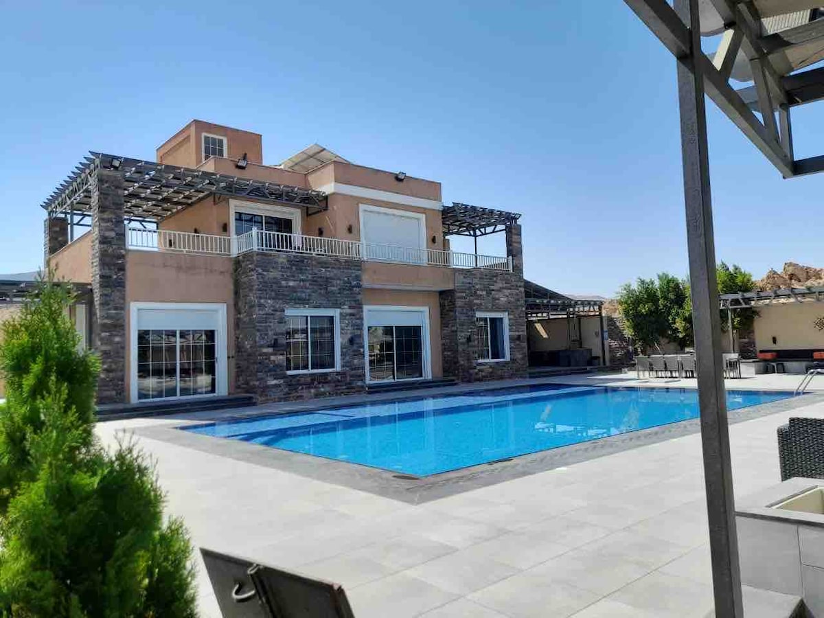 The villa's exterior is showcased, featuring a spacious layout with large glass windows. A private pool is centrally located, surrounded by a tiled deck. A shaded outdoor dining area and chairs are visible, offering a place for relaxation and enjoyment.