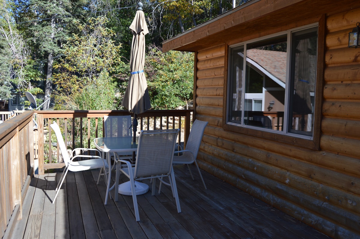A wooden deck is equipped with a glass-top table and four chairs, offering an outdoor dining space. A large umbrella provides shade, while surrounding greenery enhances the natural setting. The warm wood tones of the cabin complement the inviting atmosphere.