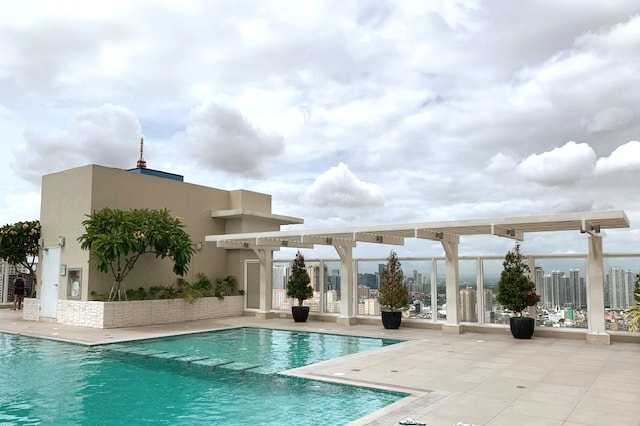 A rooftop pool area is showcased, featuring a bright turquoise water body surrounded by beige and green landscaping. Potted plants add greenery, while the skyline of Metro Manila provides a spectacular backdrop under a cloudy sky.
