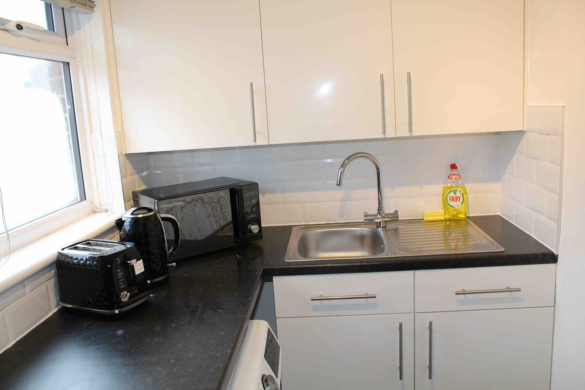 A modern kitchen is showcased, featuring sleek white cabinetry and a clean black countertop. Essential appliances, including a kettle, microwave, and dish soap are visible. A stainless steel sink with a chrome faucet is positioned under a window, allowing for natural light. The space is designed for practical daily use.
