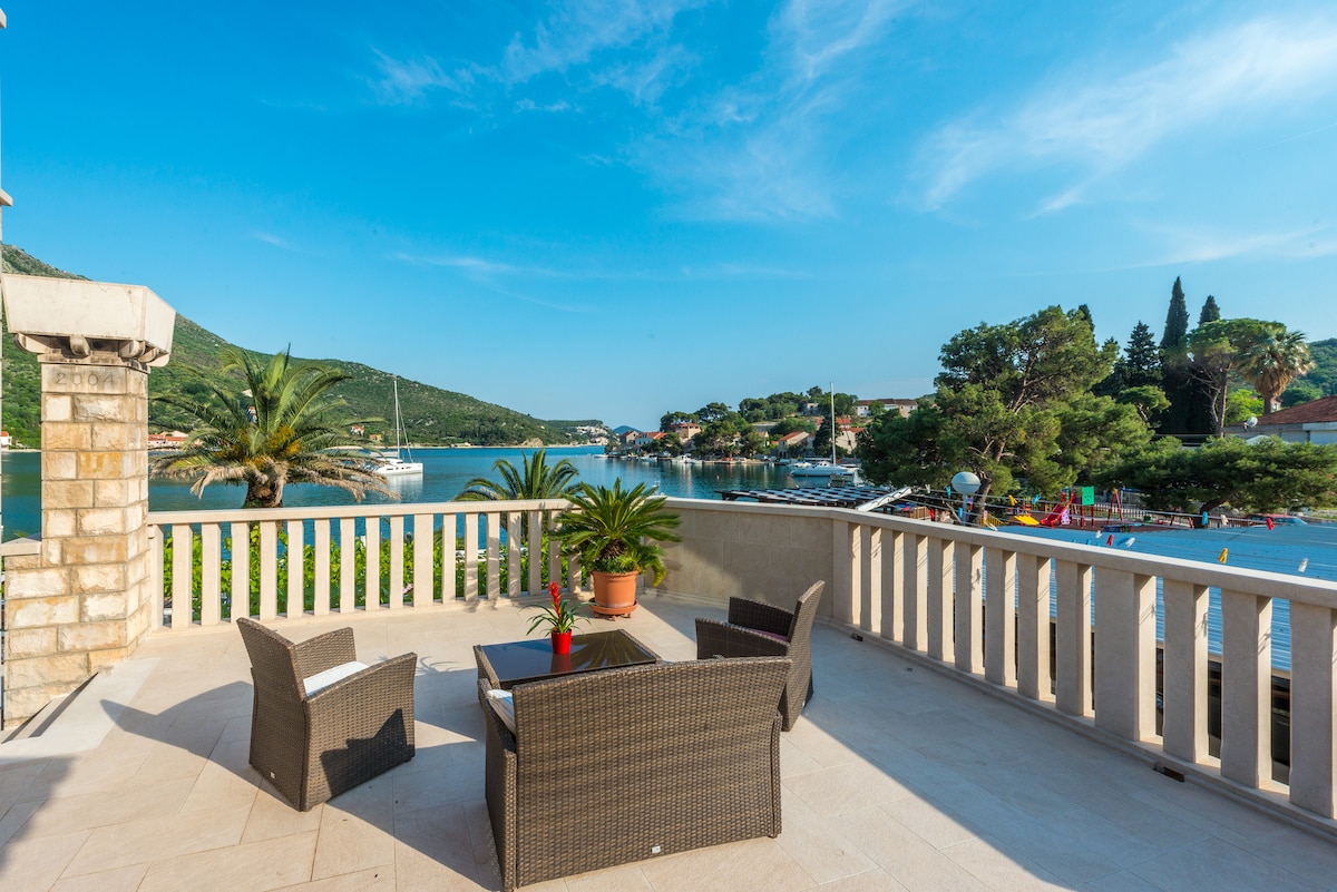 A spacious terrace features a comfortable seating arrangement with a table and chairs, surrounded by lush greenery and palm trees. The view encompasses a serene bay, dotted with boats, mountains in the distance, and vibrant, colorful houses along the shoreline.