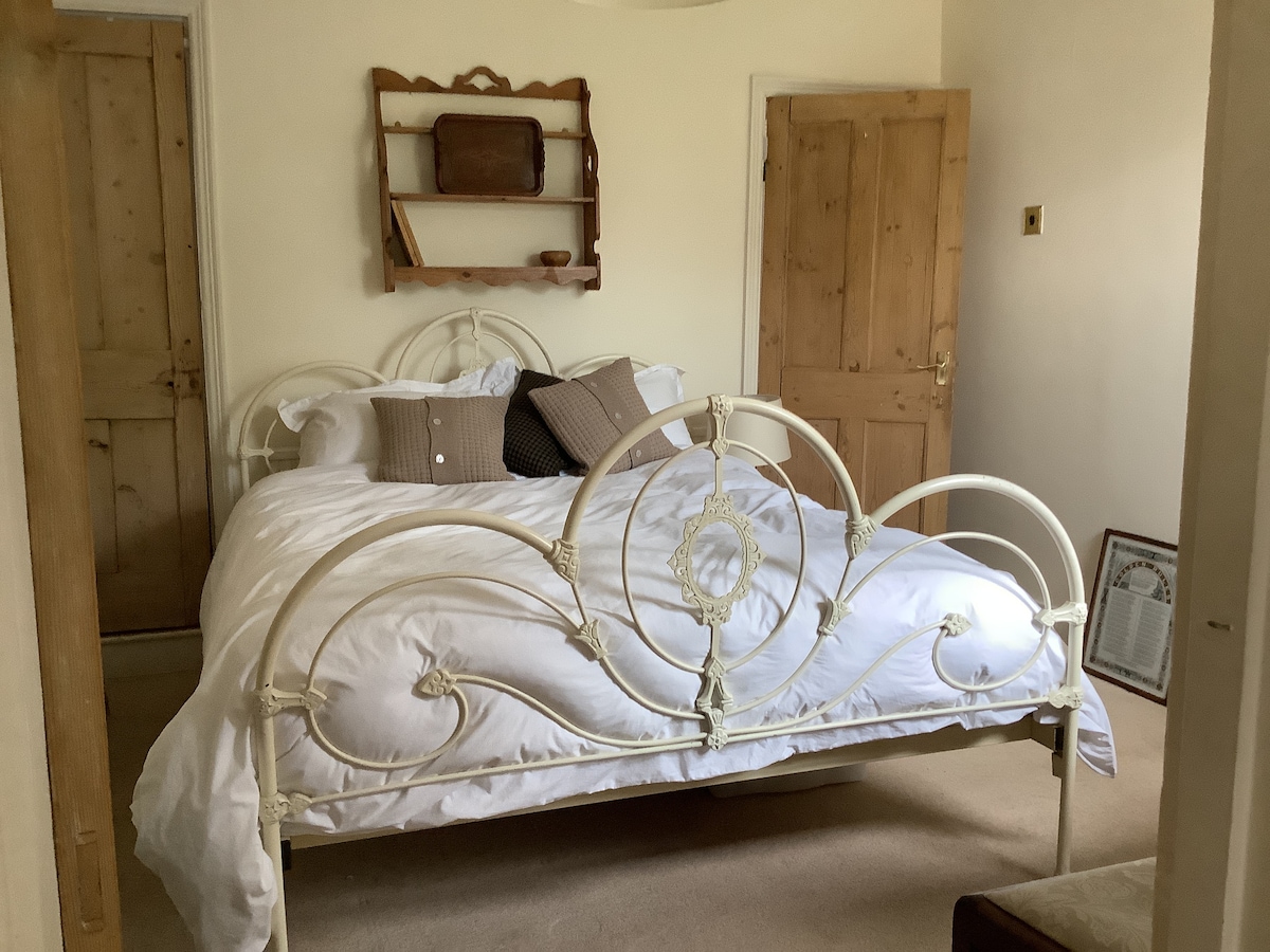 A cozy bedroom features a king-size bed with white bedding and decorative pillows. Light-colored wooden doors provide access to the room. A wooden shelf holds a few items, enhancing the room's charm. Soft lighting contributes to a relaxed atmosphere.