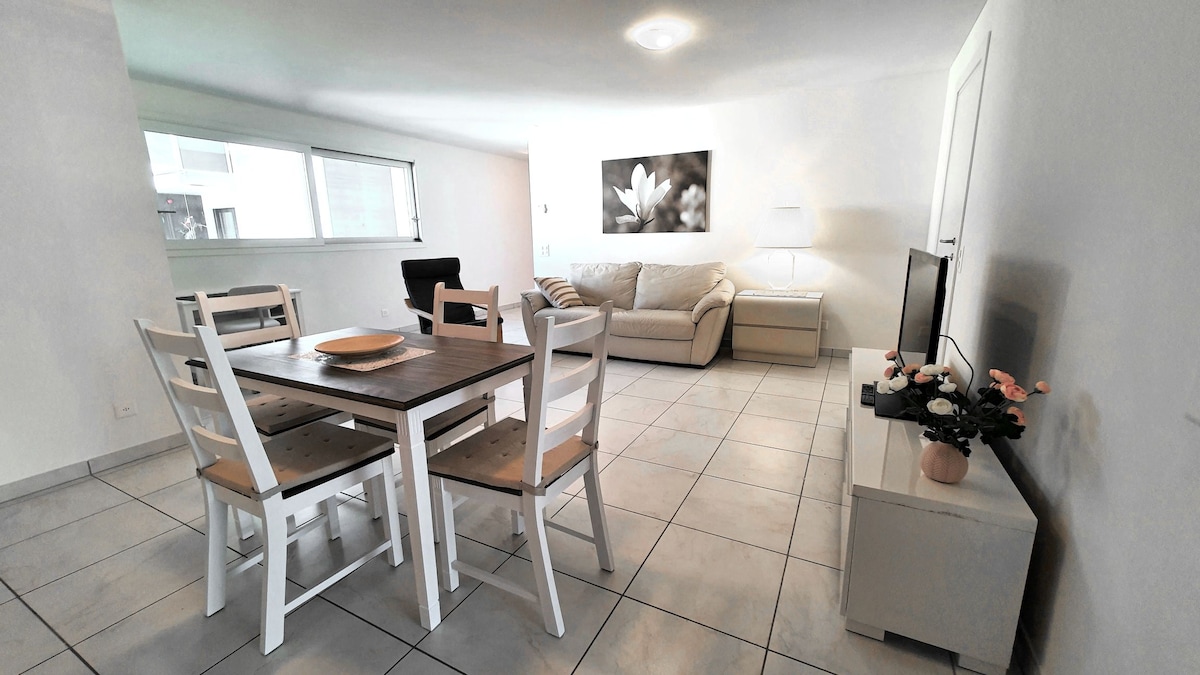 A light-filled living and dining area features a simple wooden dining table with four white chairs. A comfortable beige sofa is positioned across from a television unit, while a plant decoration adds a touch of color to the space. Large windows provide ample natural light.