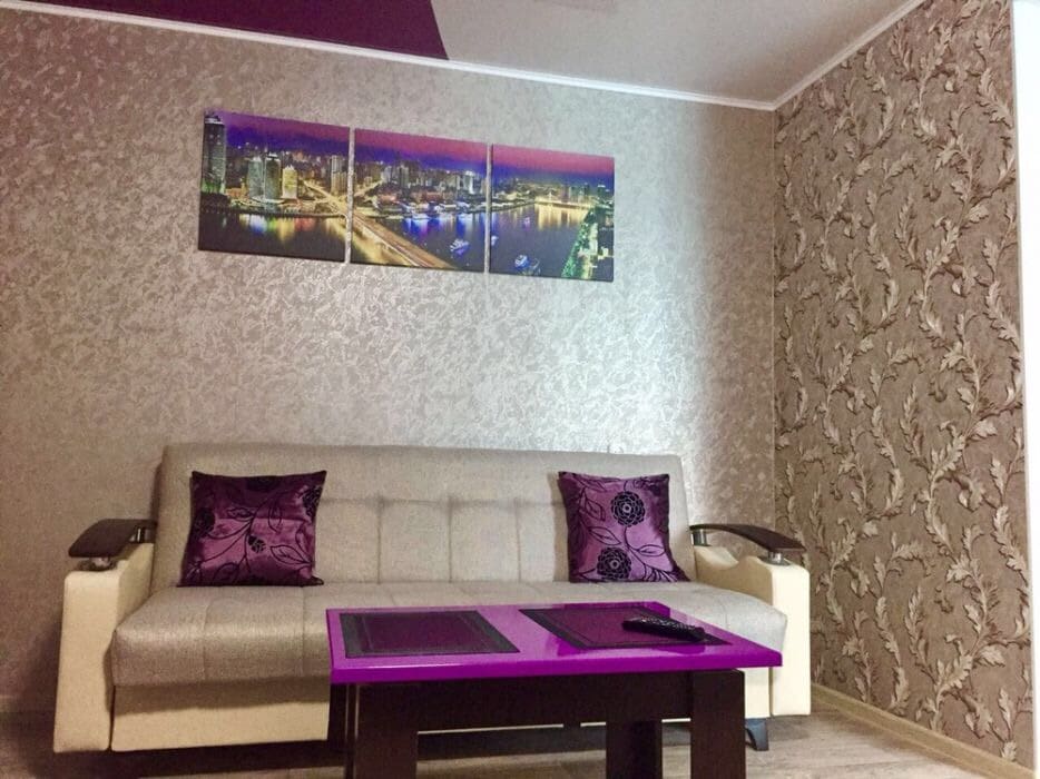 A modern living area features a light gray sofa adorned with purple accent pillows. A purple coffee table sits in front, accompanied by a remote control. The wall is decorated with a panoramic cityscape artwork, contrasting with the textured wallpaper.