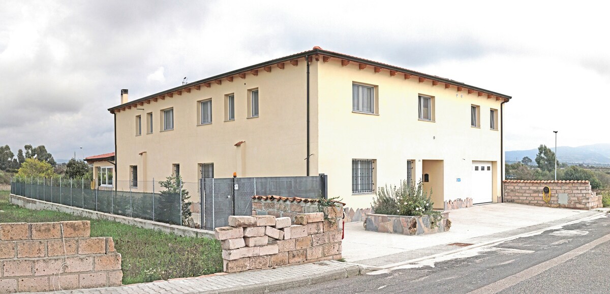 A spacious two-story structure is presented in a light beige color, featuring multiple windows and a garage. The building is surrounded by a low stone wall and a green landscape, contributing to a serene environment.