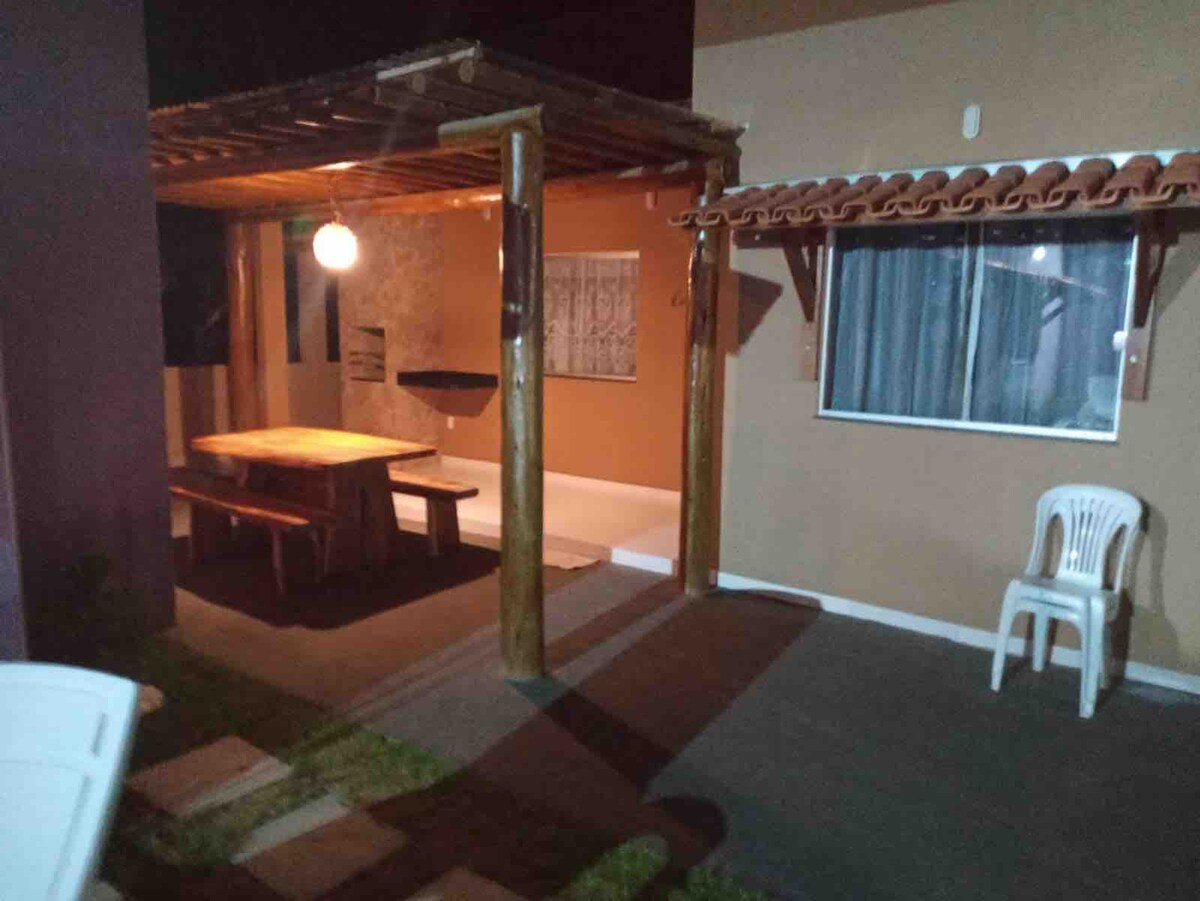 An outdoor dining area is featured under a wooden pergola, with a large table designed for several guests. A single white chair is positioned beside the table. Dim lighting enhances the atmosphere, while a window reveals a glimpse of the interior space.