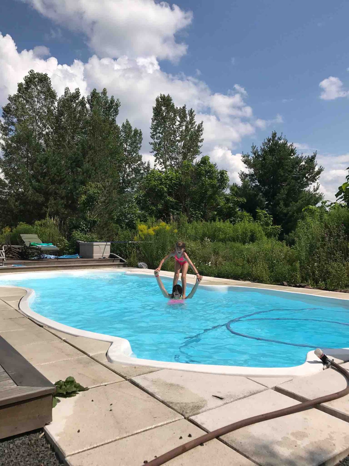 A serene swimming pool is surrounded by lush greenery, with trees framing the background. Sunlight reflects off the water's surface. Two children are playfully interacting in the pool, while sun loungers are positioned nearby on a textured paved area.