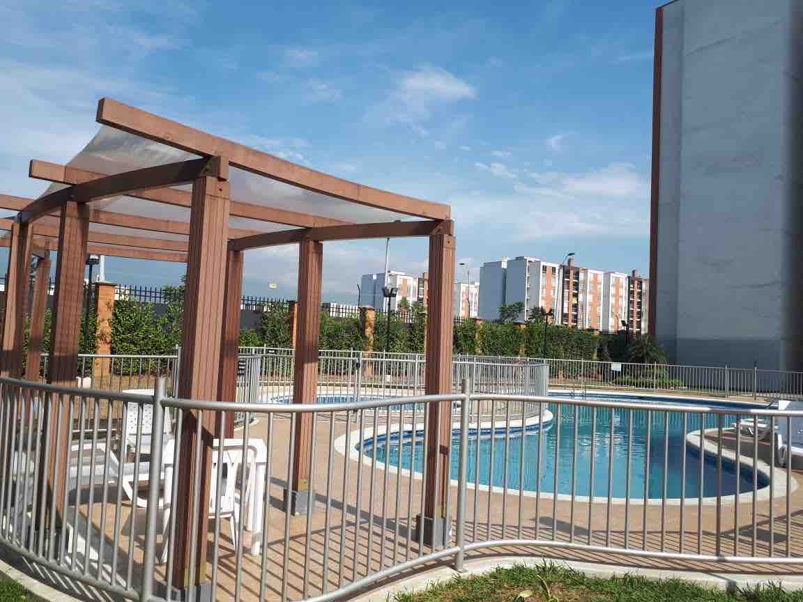 A refreshing swimming pool is enclosed by a wooden pergola and surrounded by a safety railing. Lounge chairs are arranged near the water, providing a space for relaxation. The tranquil greenery and the blue sky create an inviting atmosphere in the residential setting.