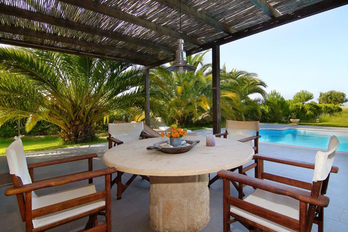 A shaded outdoor dining area is presented, featuring a round stone table surrounded by four wooden chairs. Lush green palm trees create a natural backdrop, while a private pool is visible in the distance, contributing to a serene outdoor setting.