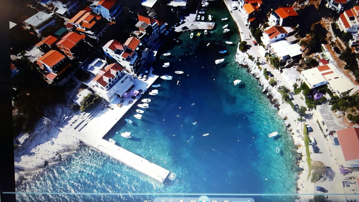 An aerial view captures a vibrant coastal village featuring white sandy beaches and a marina filled with various boats. Colorful rooftops line the waterfront, while clear turquoise waters reflect the surrounding landscape.