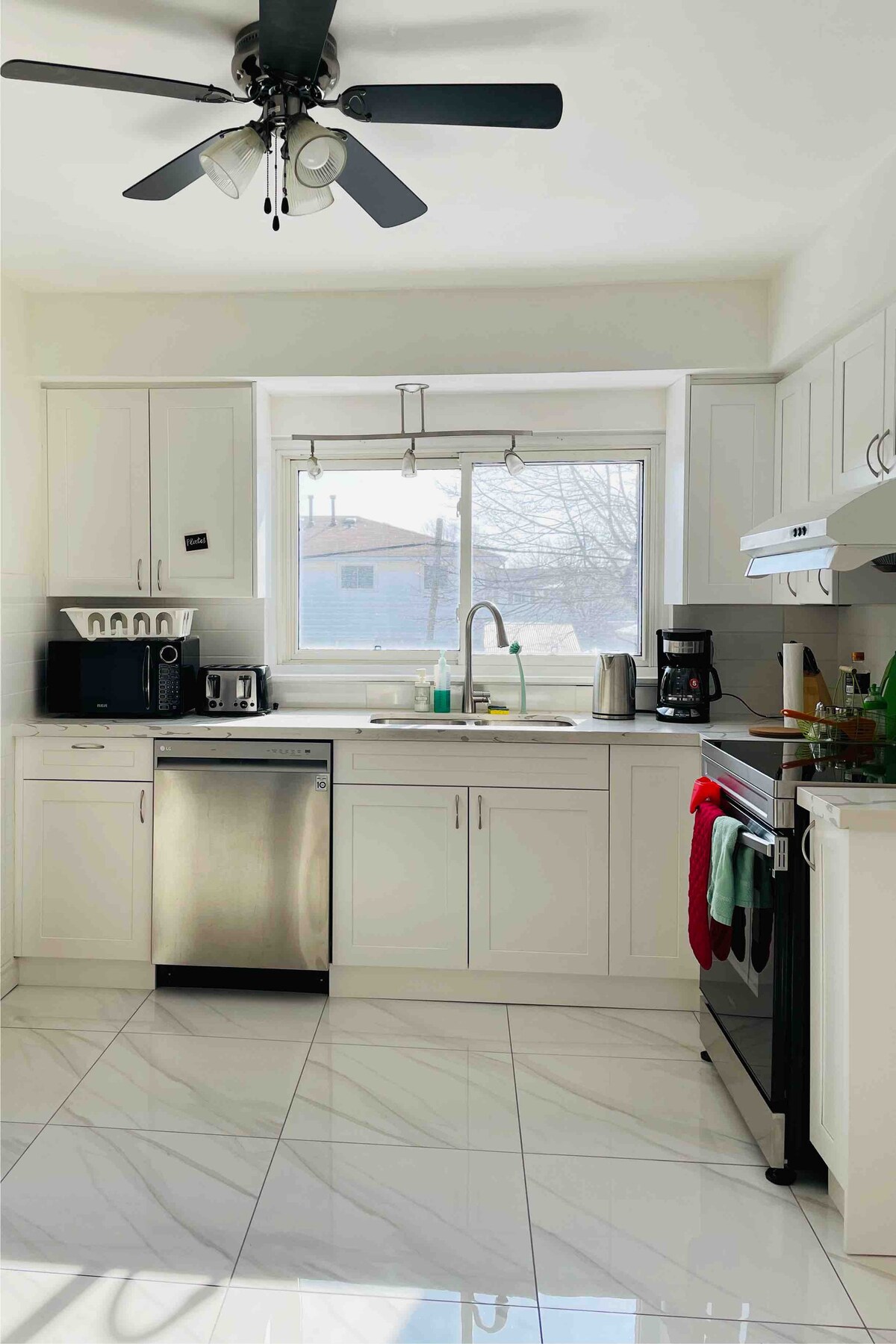A bright, fully equipped kitchen features white cabinets and stainless steel appliances. Natural light streams through a large window above the sink. Essential appliances include a dishwasher, coffee maker, and microwave, complemented by a ceiling fan and marble-style flooring.