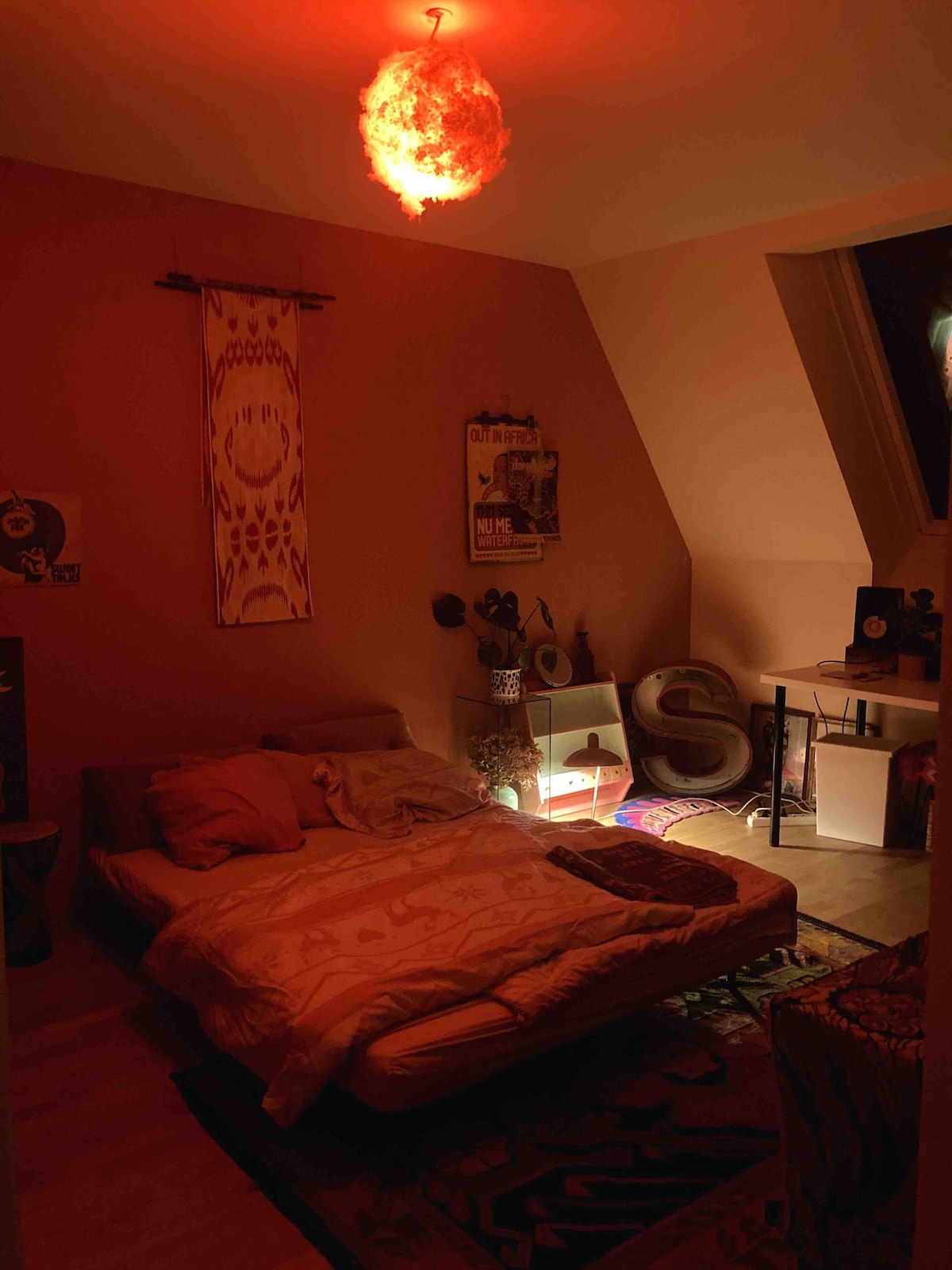 A cozy bedroom is lit with a warm red glow from a unique light fixture. The room features a spacious bed adorned with soft bedding, and decorative elements including a wall hanging, a plant, and artistic decor. A shelf displays various personal items, enhancing the artistic vibe.