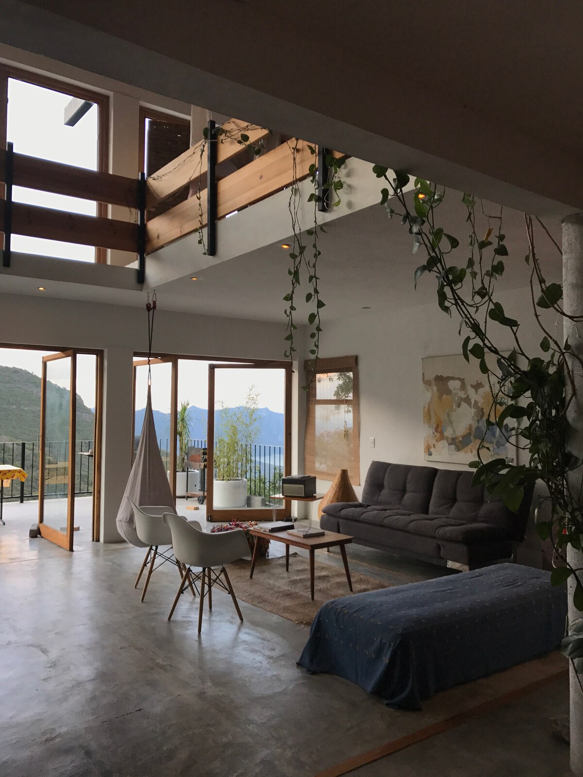 An open living area showcases a comfortable gray sofa and a round table surrounded by white chairs. Large glass doors offer views of the outdoor space and mountains. Natural light filters in, enhancing the airy atmosphere, complemented by hanging plants.