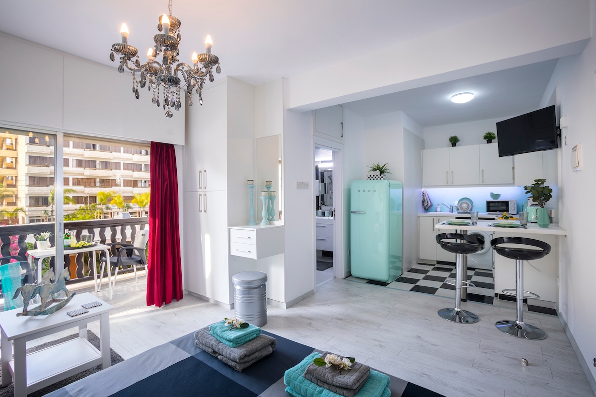 The studio apartment features a sleek modern design with a bright and airy atmosphere. A stylish chandelier hangs from the ceiling, and a kitchenette with a retro Smeg refrigerator is visible. The living area includes a cozy seating arrangement and a large window with access to a balcony.