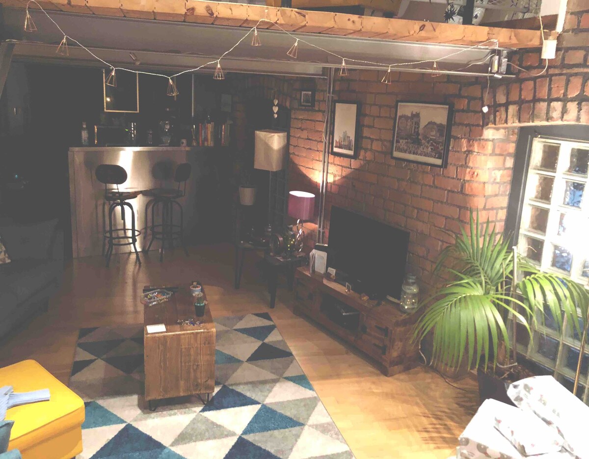 The open-plan living area features a mix of exposed brick and wooden beams, enhancing the modern yet rustic charm. A comfortable couch is positioned beside a colorful area rug, while a TV and coffee table sit in front. Subtle lighting is provided by lamps and string lights.