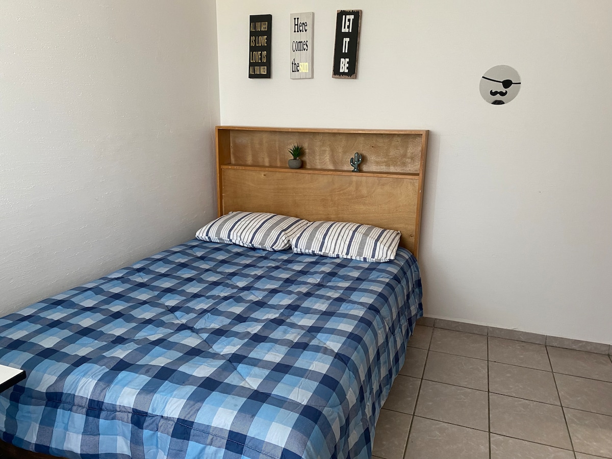 A comfortable sleeping area features a spacious bed with a plaid blue blanket. Above the bed, small decorative plants are placed on a wooden headboard. Simple art pieces adorn the wall, contributing to a relaxed ambiance.