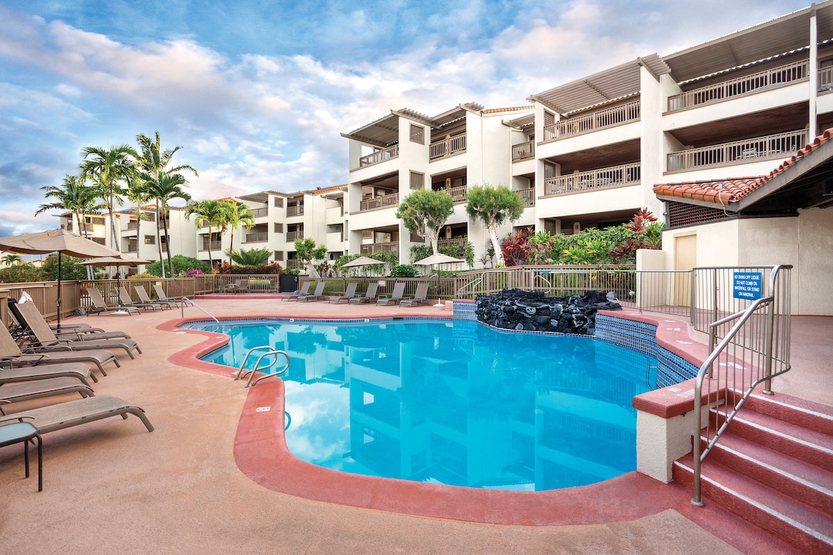 A resort pool area is visible with clear blue water and a rock feature. Lounge chairs are arranged around the pool, and palm trees are scattered throughout the landscape. The two-story building provides a backdrop, featuring multiple balconies and flower gardens.