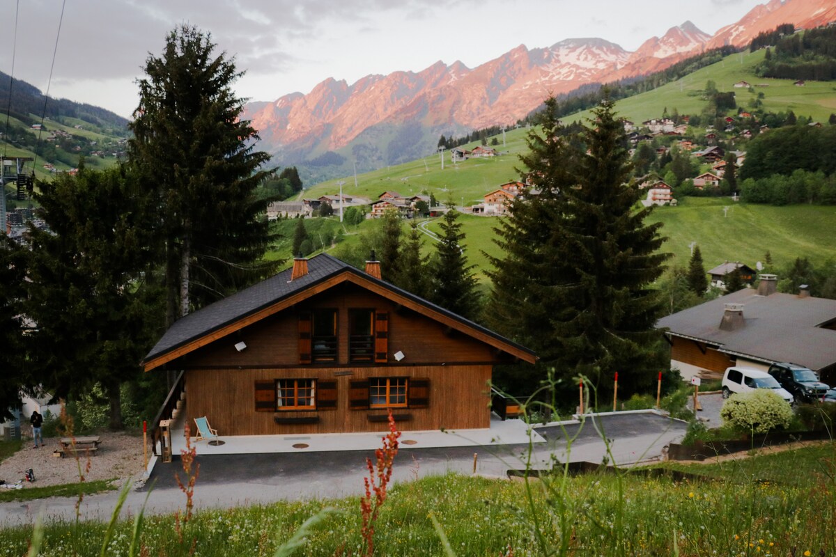 The exterior of a wooden chalet is set amidst green grass and trees, with a stunning mountain backdrop. The building features a large balcony and a spacious parking area. The landscape showcases rolling hills and peaks, providing a serene alpine environment.