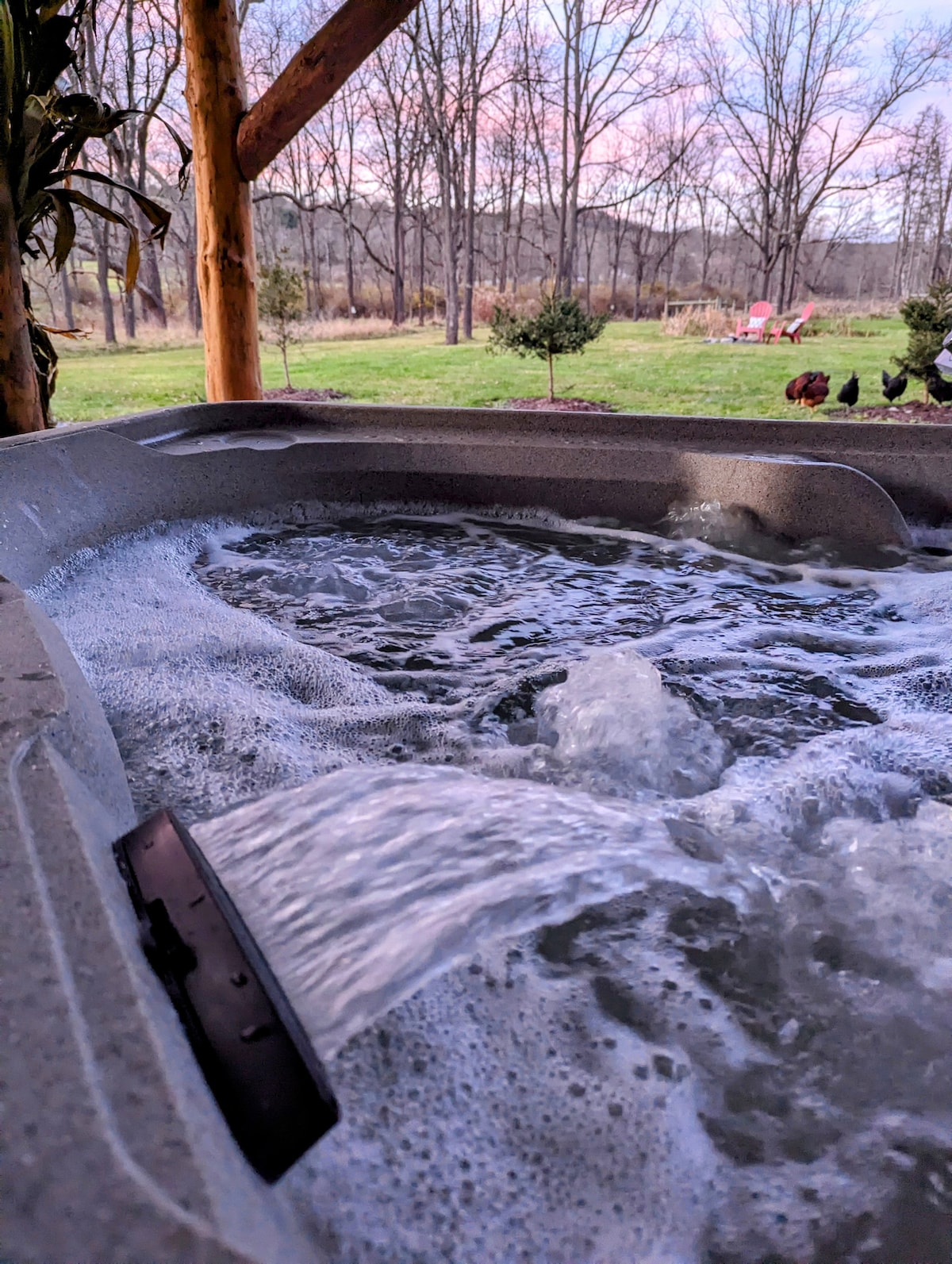 A hot tub is shown with bubbling water, nestled on a patio covered by a wooden structure. In the background, a grassy area is visible with scattered trees and two Adirondack chairs, creating a peaceful outdoor setting.