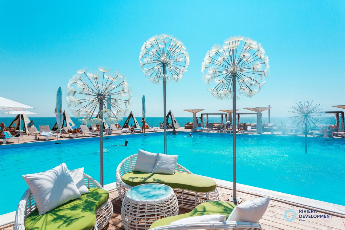 A modern pool area features a large turquoise pool surrounded by comfortable lounge chairs and umbrellas. Decorative elements resembling oversized dandelions add a playful touch, and distant figures enjoy the water under clear blue skies.