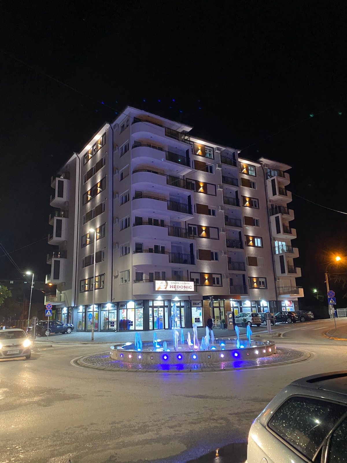 A modern multi-story building is illuminated at night, showcasing a sleek façade with balconies. A circular fountain is positioned in the foreground, featuring blue lights that reflect off the water. Surrounding streets include vehicles parked along the sides.