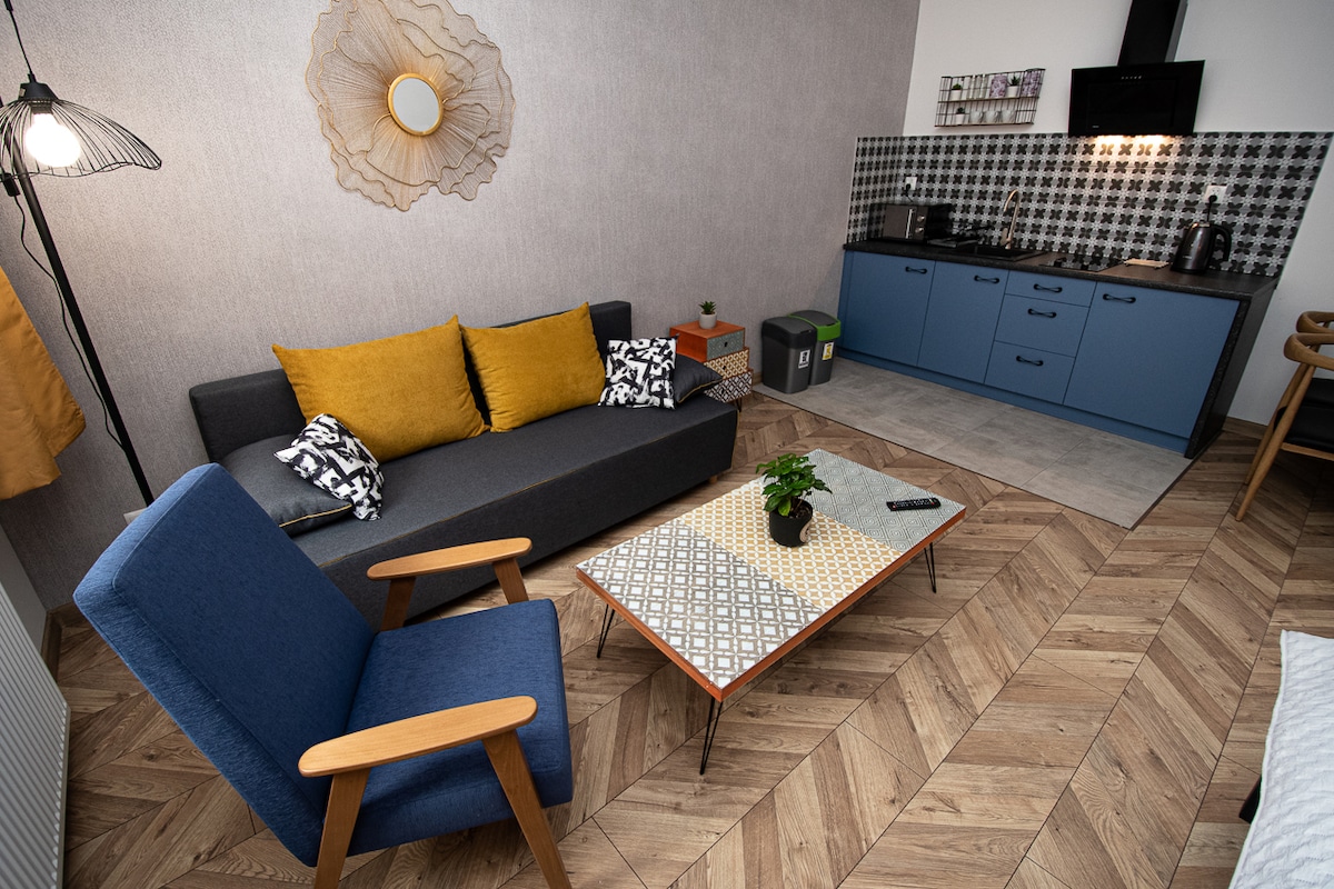 A living area features modern furnishings, including a dark gray sofa adorned with yellow pillows and a minimalist coffee table. A blue armchair adds contrast. The kitchenette has a black countertop with patterned tiles and includes kitchen appliances, creating a functional space for dining and relaxation.