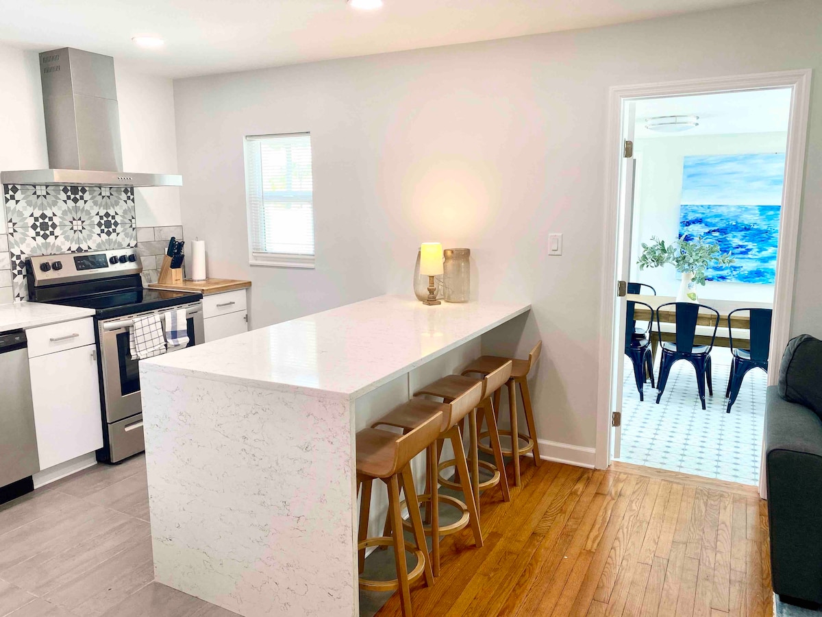 A modern kitchen features stainless steel appliances and a sleek island with seating for four. Bright mosaic tiles add a touch of design, while light streams in through a nearby window. A seamless transition to the dining area can be observed through the open doorway.