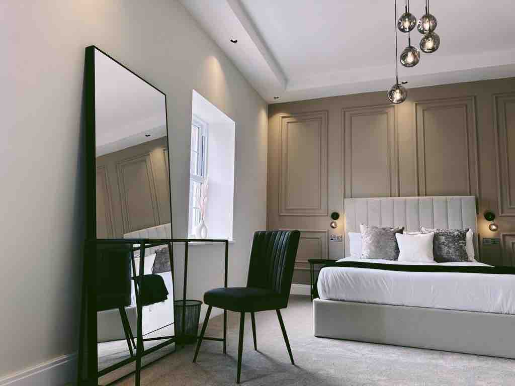 A spacious bedroom featuring a king-size bed with a soft headboard, complemented by decorative pillows. A stylish mirror and a sleek black chair are placed near a window, enhancing the room's modern design and inviting natural light.