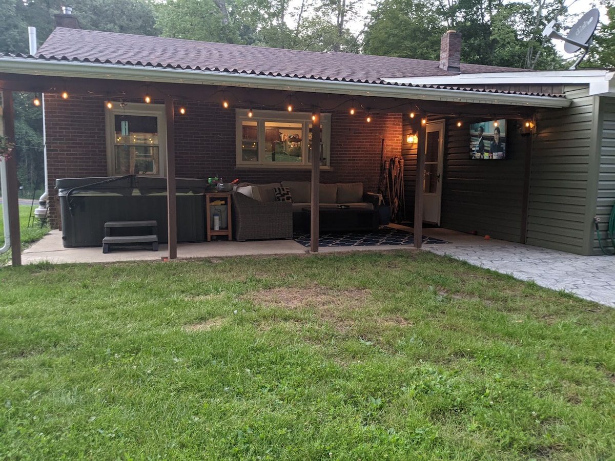 A covered patio area features comfortable seating arranged around a low table, with a hot tub nearby. Soft lighting illuminates the space, creating a cozy atmosphere. A TV is mounted on the wall, and the grassy yard extends beyond the patio, offering ample outdoor space.