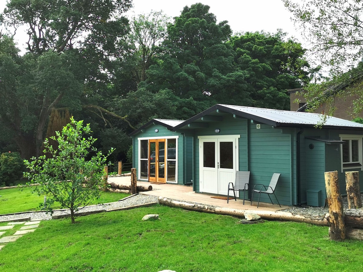 A serene log cabin is surrounded by lush greenery, featuring large glass doors and a welcoming front porch with seating. A neatly maintained lawn and stepping stones lead up to the cabin, providing a peaceful, natural setting.