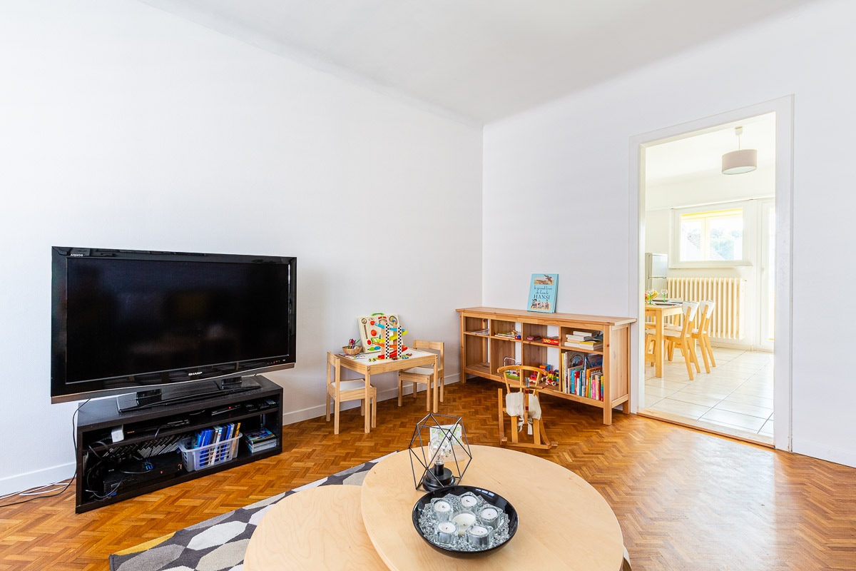 A bright living room features a large television and a low circular coffee table at the center, surrounded by light wood flooring. Two child-sized chairs are positioned beside a shelving unit filled with books and toys, with a dining area visible through an open doorway.
