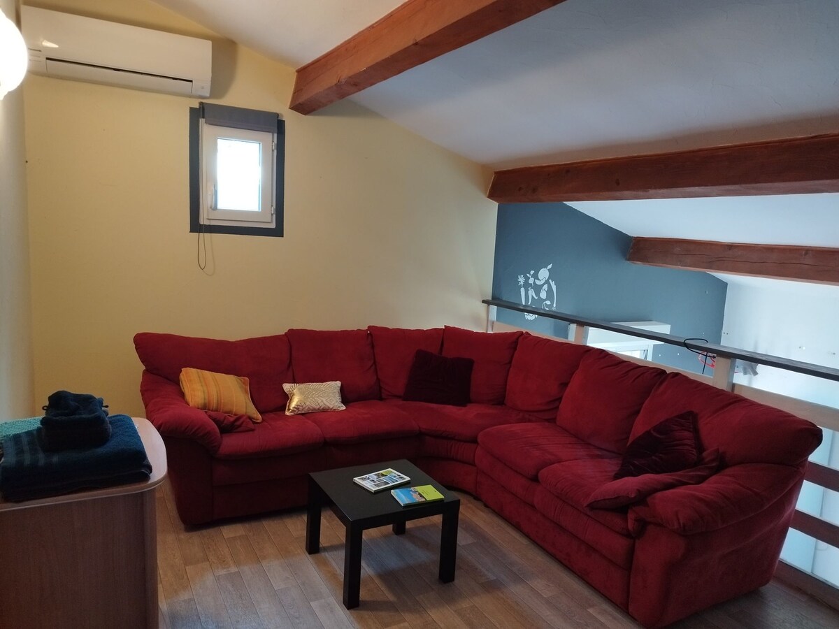 A cozy living area is furnished with a large red sectional sofa, which is complemented by decorative cushions. A small black table is positioned in front, adding functionality. A window above provides natural light, and wooden beams enhance the open feel of the space.
