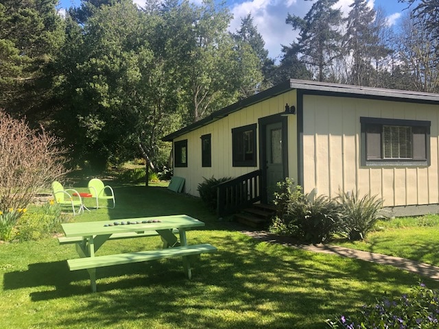 The exterior of The Shed Guesthouse is depicted, featuring a charming building with a light-colored facade surrounded by lush green grass. A picnic table is positioned in front, while colorful chairs are arranged nearby, inviting outdoor relaxation. Trees provide shade, enhancing the serene atmosphere.