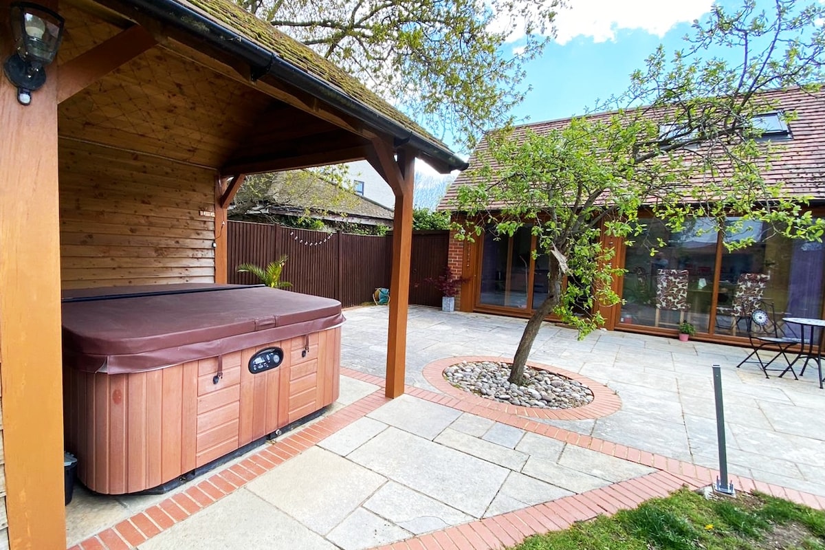 A private patio area features a luxurious hot tub nestled under a wooden structure. A circular stone arrangement surrounds a small tree, while paved stones create a spacious surface. Large glass doors provide access to the retreat, showcasing natural light and outdoor views.