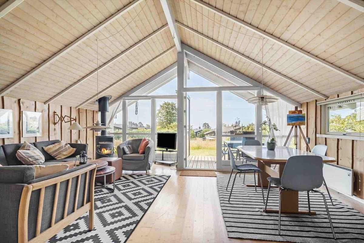 The open living space features a high, angled wooden ceiling, with large glass doors providing natural light and garden views. A cozy seating area includes a dark sofa and armchair, accompanied by a dining table with modern chairs. Decorative accents enhance the inviting ambiance.