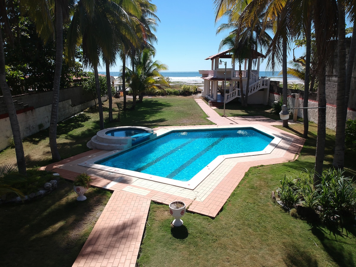 A clear view of a private pool area featuring a rectangular swimming pool and a circular jacuzzi, surrounded by manicured grass and palm trees. The beach and ocean are visible in the background under a bright blue sky, creating a serene outdoor setting.