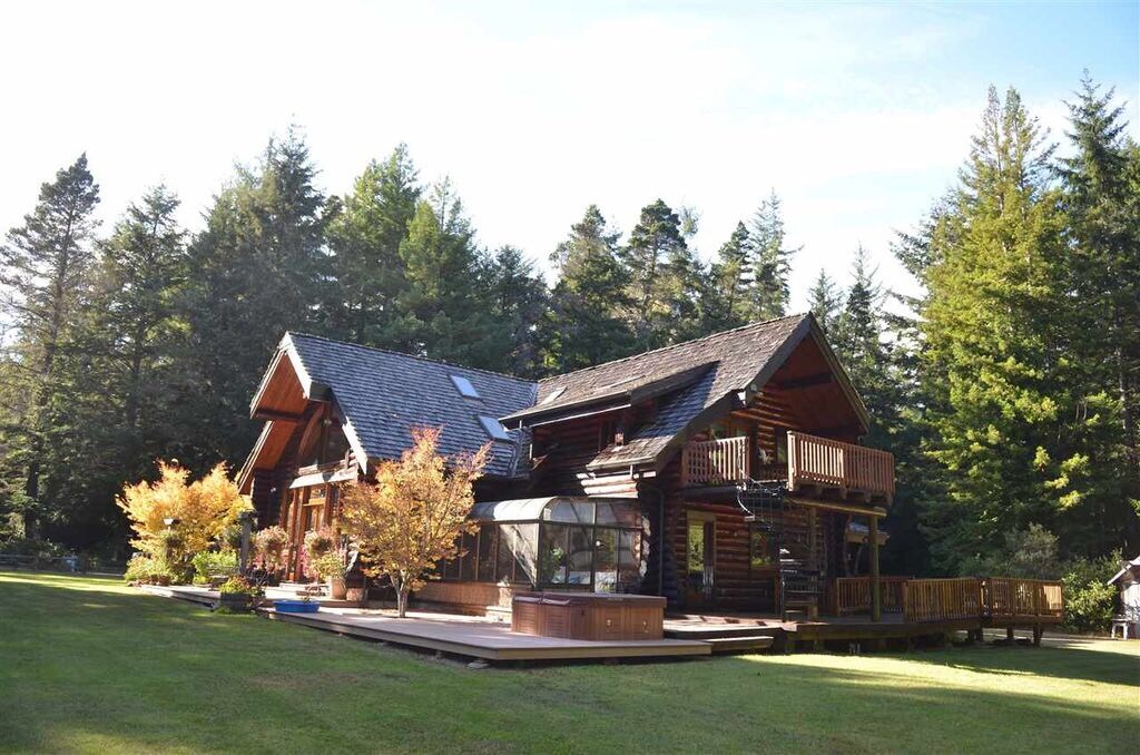 A rustic log cabin is surrounded by lush forest, featuring a large deck with seating and a hot tub. A well-maintained lawn complements the natural landscape, while trees provide a serene backdrop. Skylights and balconies enhance the cabin's charm.
