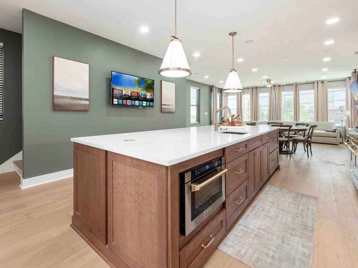 A modern kitchen is highlighted by a large island with a white countertop and wooden cabinetry. A built-in microwave is integrated, while a wall-mounted television is visible. Natural light fills the space through multiple large windows, complemented by a neutral-colored rug.