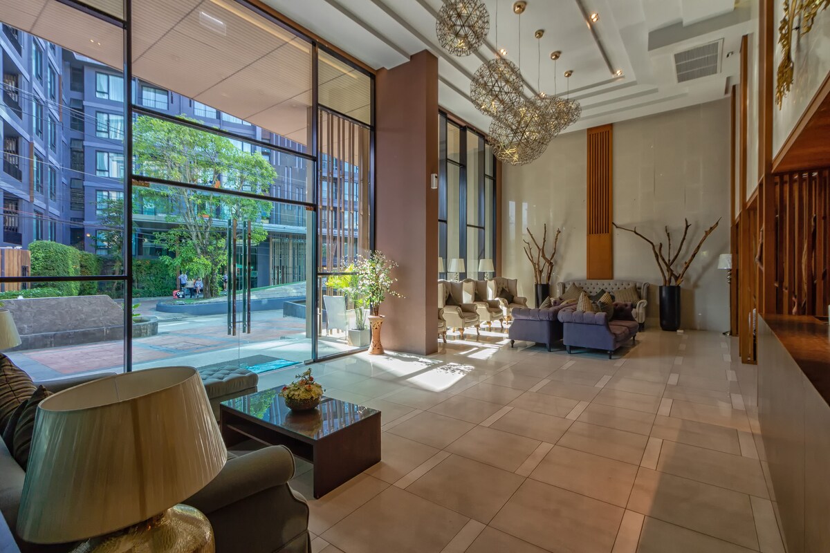 The spacious lobby features large glass windows, allowing natural light to fill the area. Comfortable seating arrangements are provided with stylish sofas and a coffee table. Elegant light fixtures hang from the ceiling, enhancing the modern design alongside decorative plants.