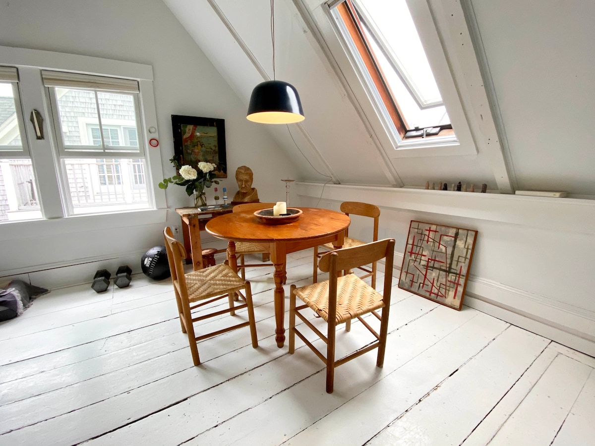 A small dining area is visible featuring a round wooden table surrounded by four wooden chairs. A pendant light hangs above, and natural light filters through a skylight. Floral arrangements and artwork are displayed around the space, enhancing its cozy ambiance.