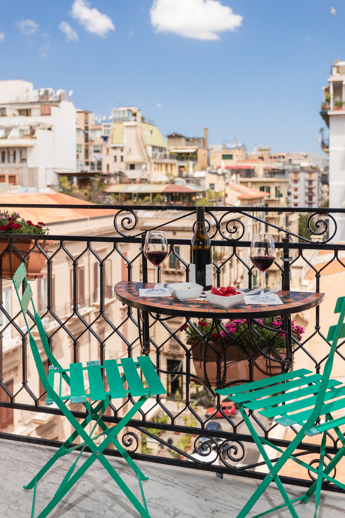 A balcony scene features a small round table set with two glasses of red wine and a plate of strawberries. Green metal chairs are positioned around the table, with vibrant flower pots adding color. A view of the city skyline is visible in the background.