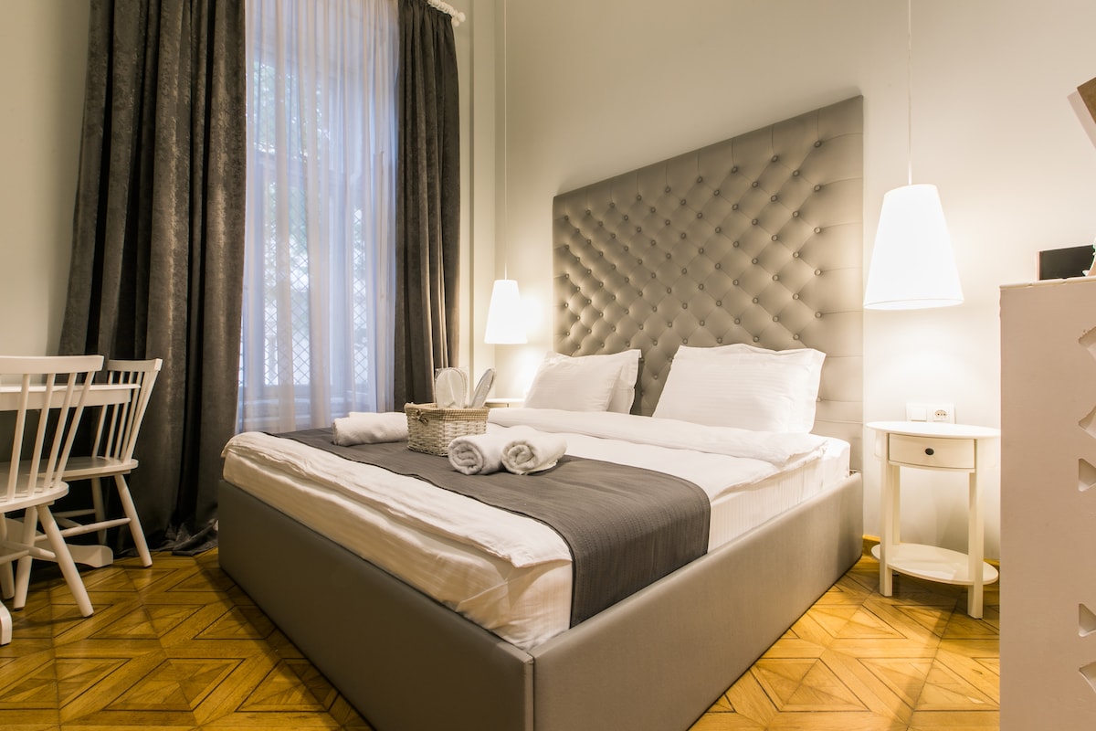 A spacious sleeping area features a large, upholstered bed adorned with crisp linens and two accent pillows. Soft lighting from pendant fixtures illuminates the room, complemented by tall windows dressed with sheer curtains. A small dining table and chairs are positioned at one side.
