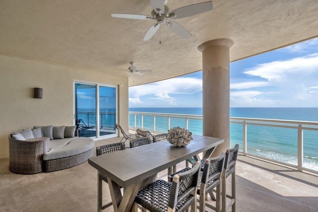 A spacious oceanfront balcony features a large dining table surrounded by six woven chairs. A cozy circular lounge area with plush cushions is positioned nearby, while ceiling fans provide a cooling breeze. The ocean view is unobstructed, offering a serene backdrop to the gathering space.