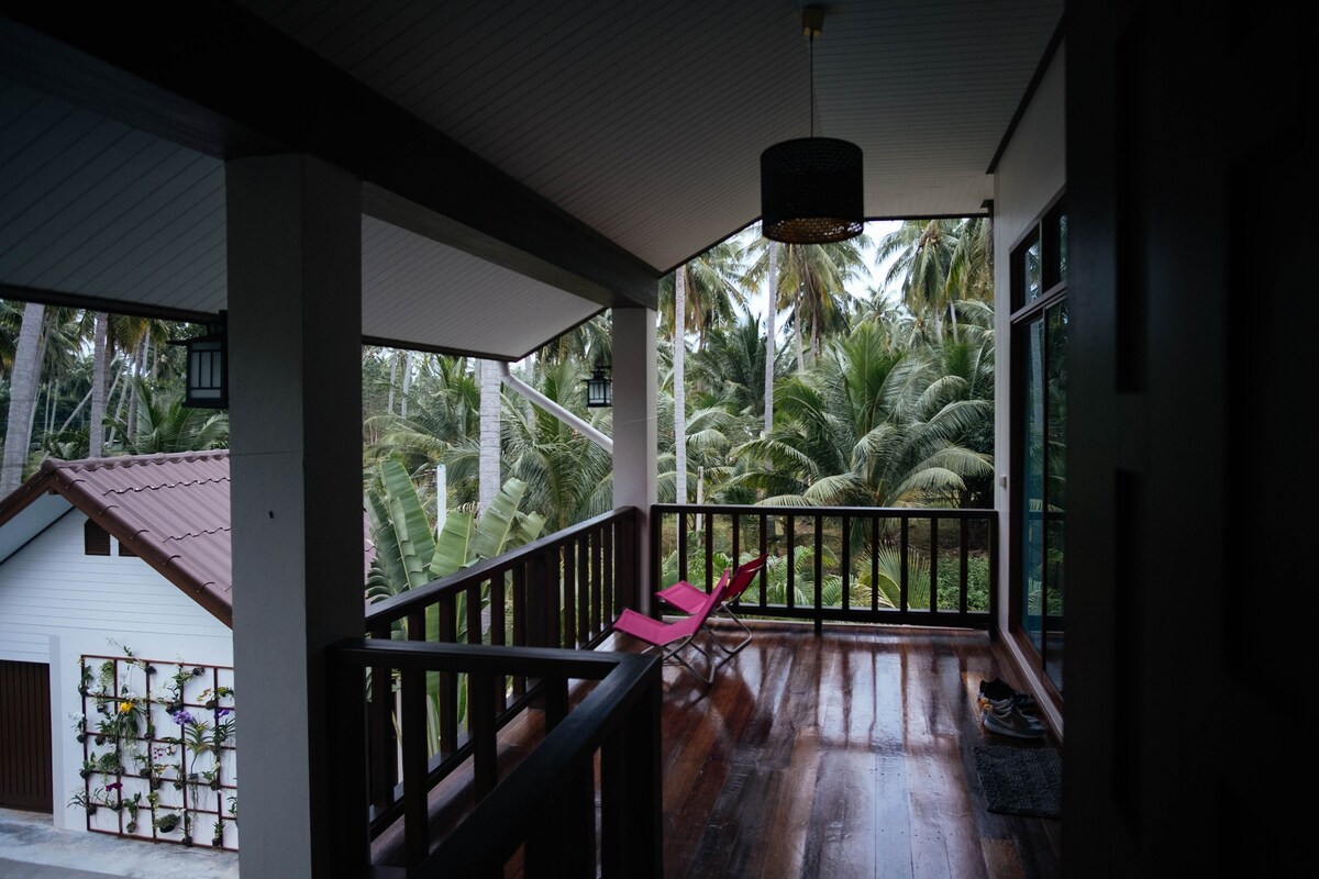 A spacious balcony is framed by wooden railings, offering views of lush greenery. Two comfortable lounge chairs are positioned for relaxation, with soft lighting provided by an overhead pendant lamp. The natural light enhances the warm tones of the polished wooden floor.