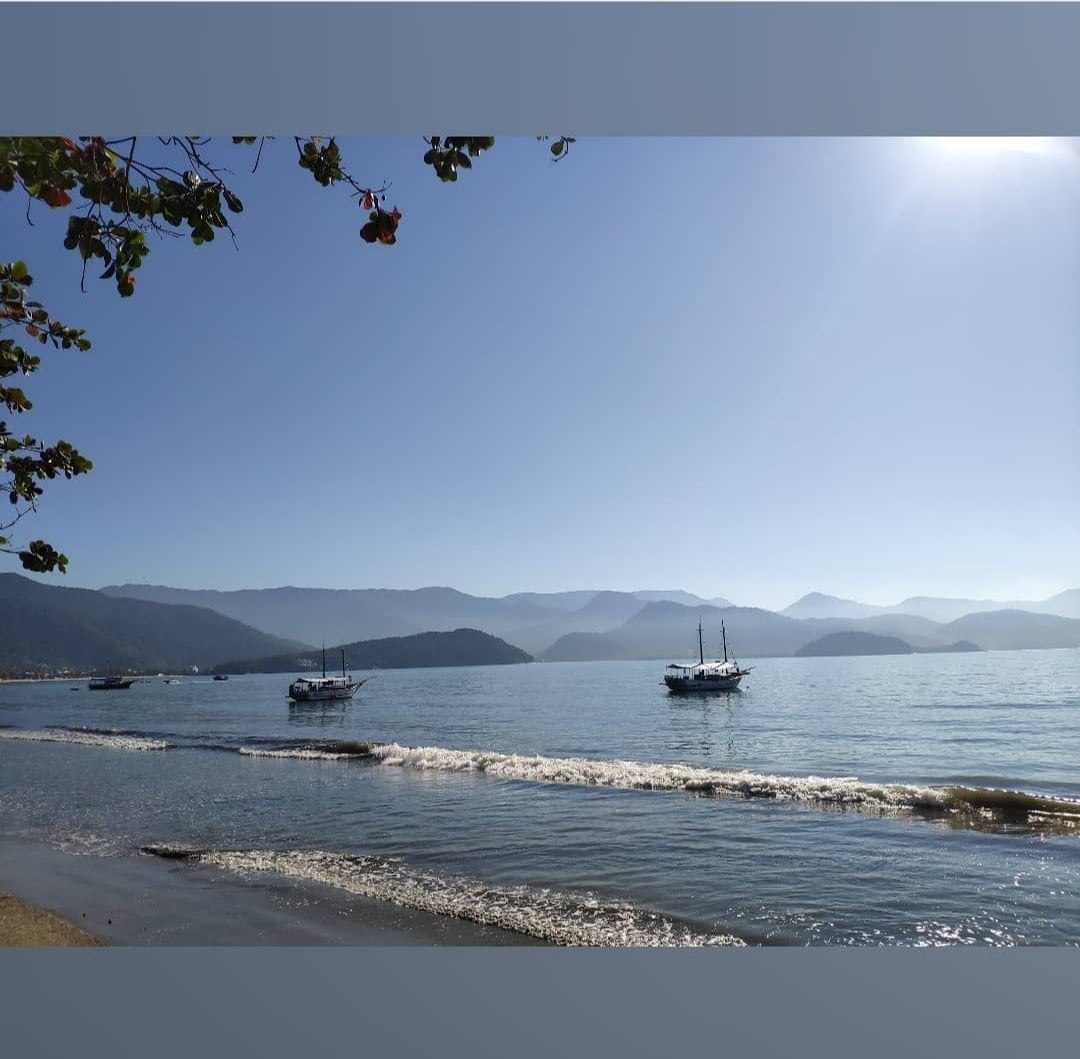 Two boats are anchored on calm waters, gently reflecting the light from the clear sky. The shoreline features a sandy beach, with distant mountains providing a serene backdrop. Soft waves can be seen lapping against the shore, contributing to a tranquil coastal scene.