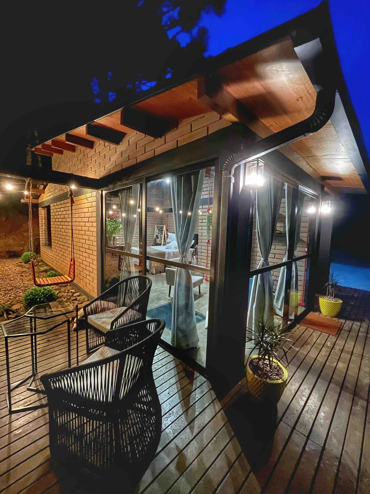 The exterior of the chalet is presented at night, illuminated by soft lighting. Large windows showcase a cozy indoor area, while a wooden deck features two black chairs and potted plants. Draped curtains add a touch of warmth to the inviting atmosphere.
