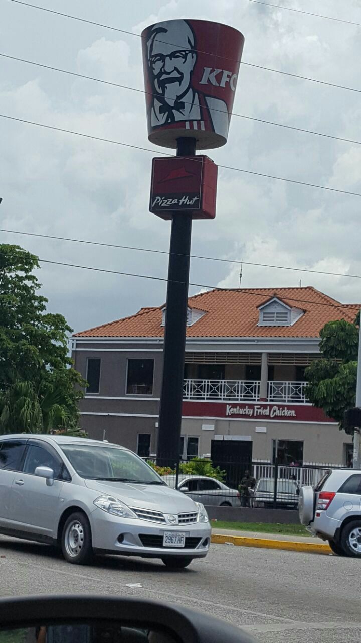 A tall sign stands prominently, featuring the recognizable KFC bucket logo at the top, above the Pizza Hut logo. A white car is parked in the foreground, with a building in the background that has a red roof and multiple windows, indicating a commercial area.