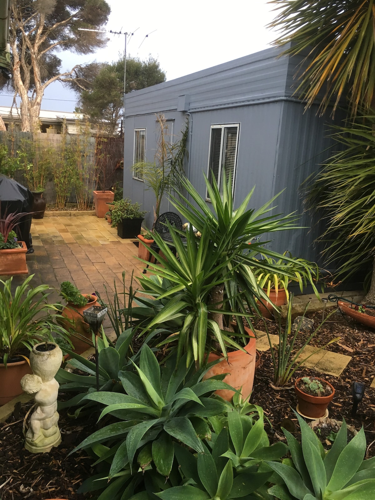 The image features a separate self-contained studio nestled within lush garden surroundings. Potted plants and greenery add a vibrant touch. A stone path leads to a private entrance, while a BBQ area is visible in the background, enhancing the outdoor space.